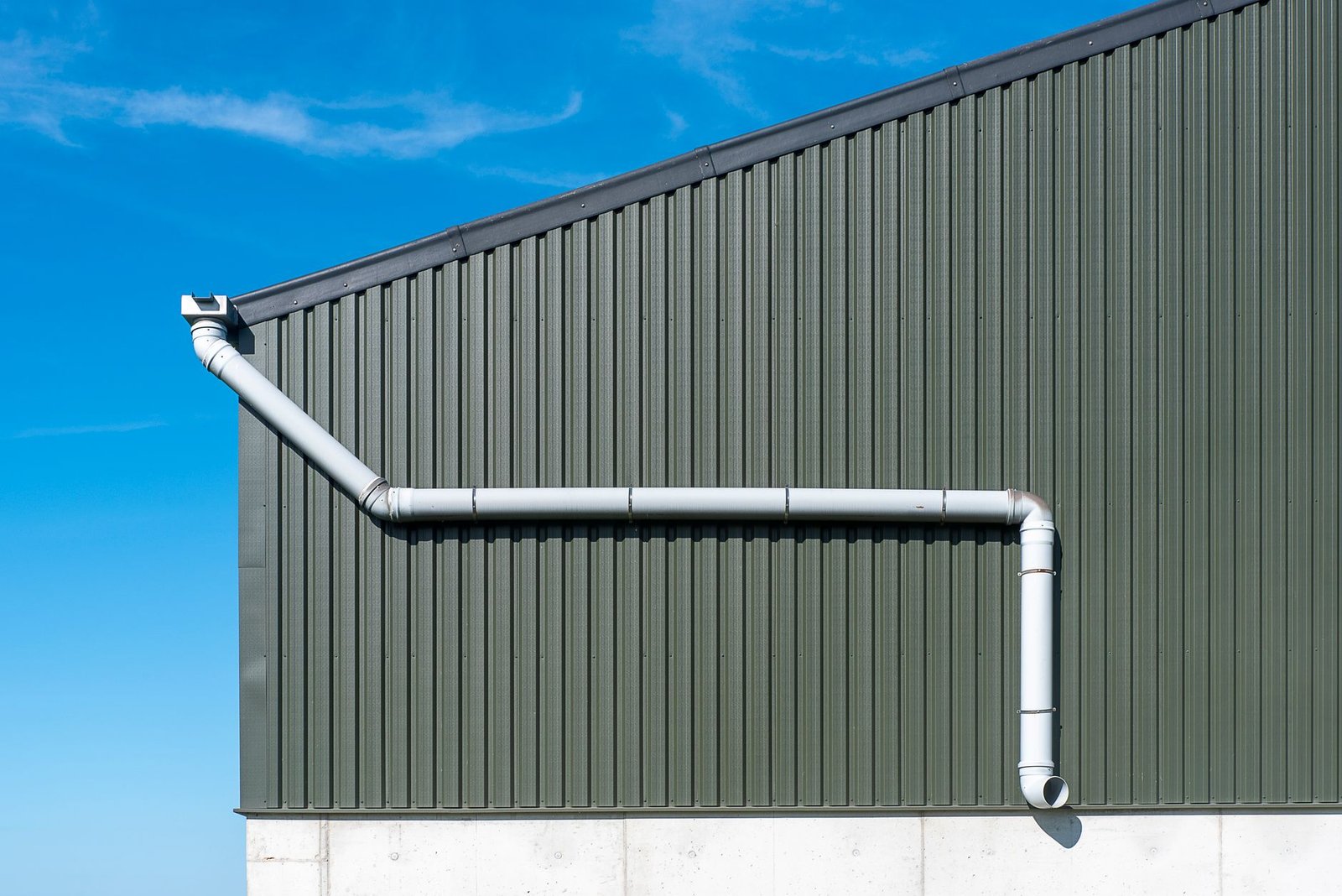 Exterior view of a corrugated metal building with a rainwater drainpipe against a clear blue sky.